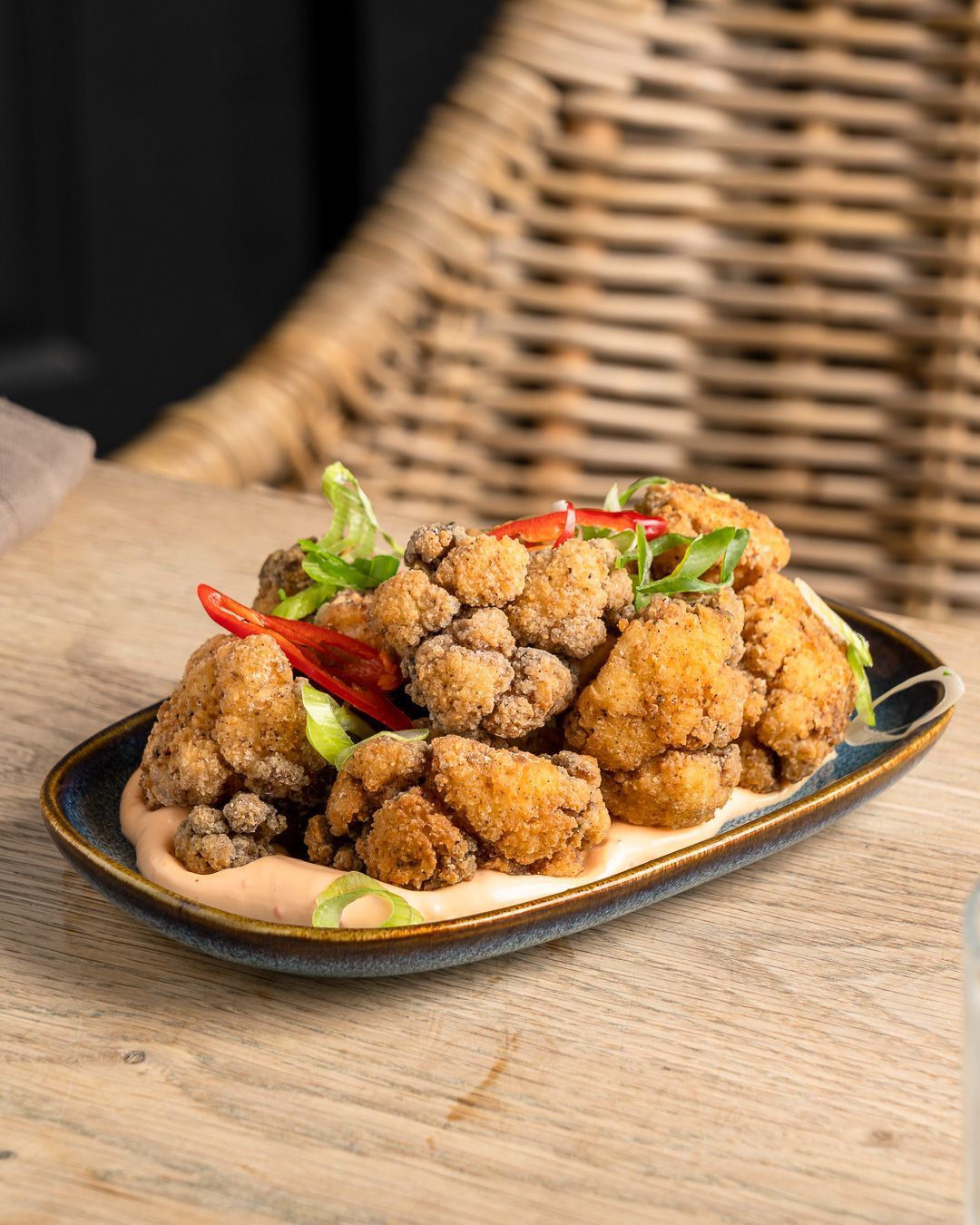 Fried cauliflower with sauce, garnished with red peppers and green onions, served on a dark plate, wood background.