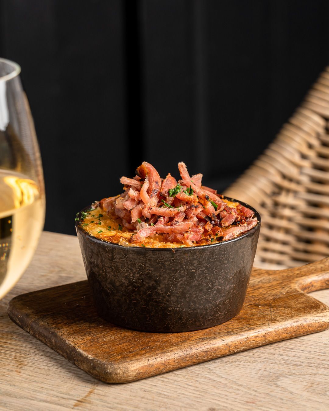 Mac and cheese topped with crispy bacon, served in a black bowl on a wooden board next to a glass of white wine.