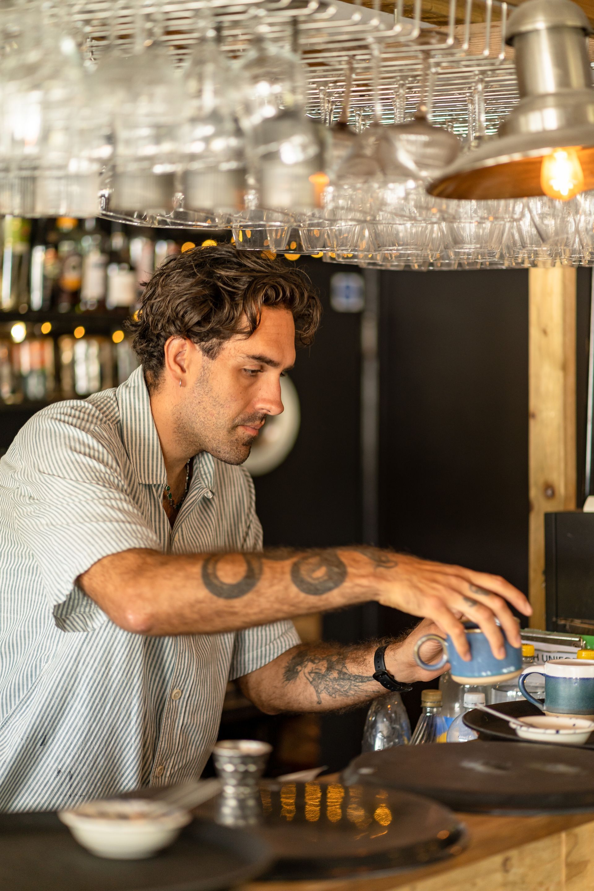 Man preparing coffee at a bar, reaching for a cup. Glasses hang above.