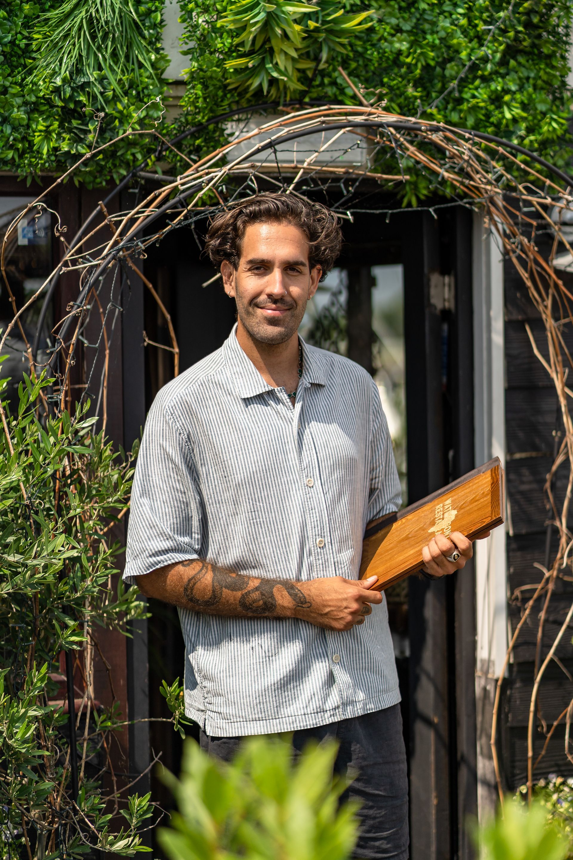 Man with curly hair holds wooden frame, stands in front of a door with archway covered in greenery.
