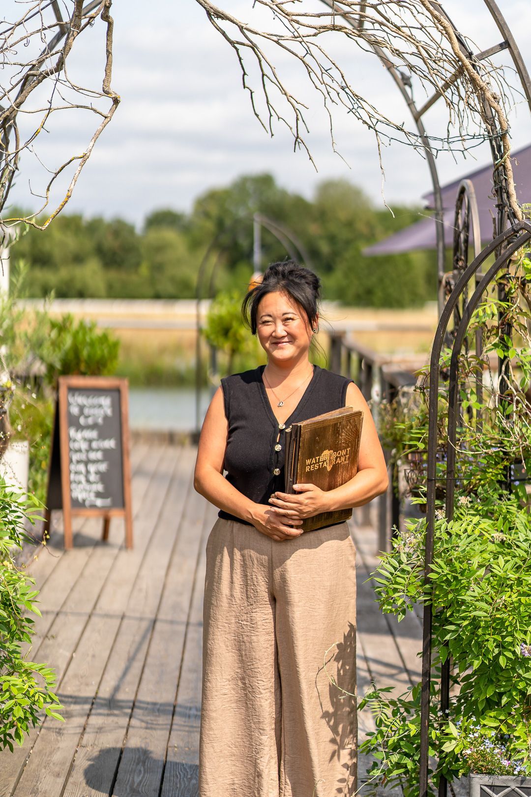 Woman holding book, standing on wooden walkway, arched trellis, outdoor setting.