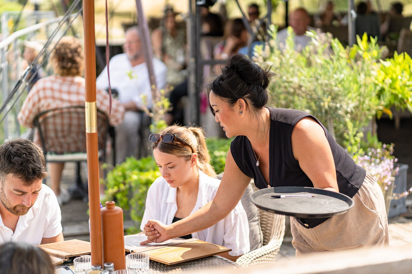 Waitress serving a table outside a restaurant. Customers seated, reviewing menus. Sunny day.