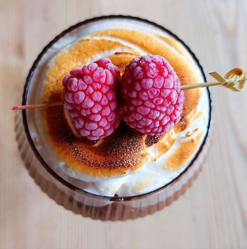 Cocktail with toasted marshmallow topping, garnished with raspberries on a skewer.