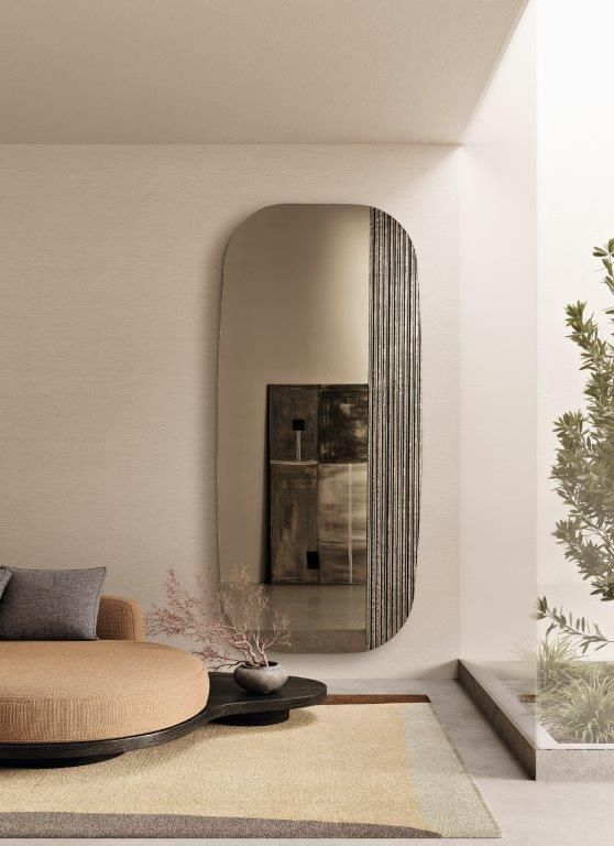 Soggiorno minimalista con specchio rotondo, divano beige, tappeto intrecciato e una pianta in vaso vicino alla finestra.