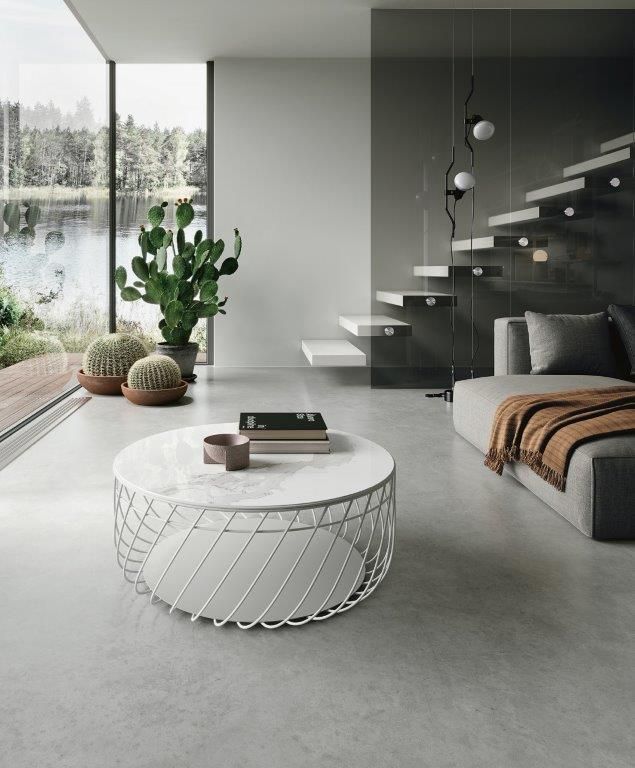 Soggiorno moderno e minimalista con vetrata a tutta altezza, divano grigio, tavolino rotondo in filo metallico e scaffalatura sulla scala.