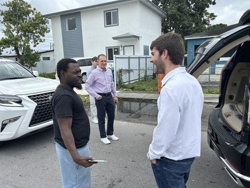 Three men standing and talking in a driveway beside parked cars and a white house.