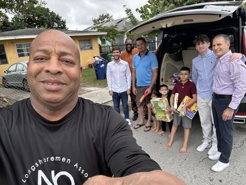 Group posing outdoors beside a black SUV; man in front takes a selfie, several people smile, two children hold gifts.
