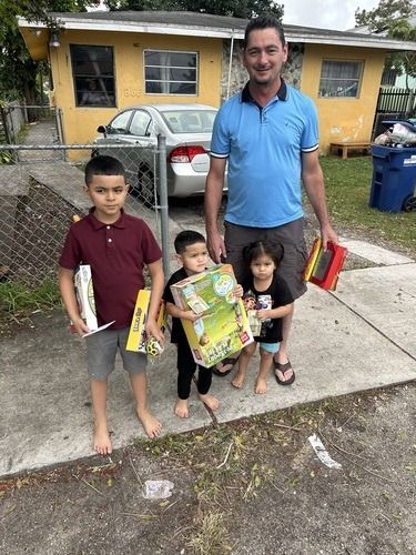 Man standing with two children holding bags on a sidewalk in front of a yellow house and parked car