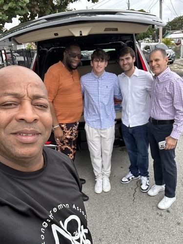 Five men posing in front of an open car trunk on a street, smiling at the camera.