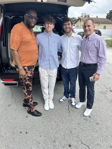 Four men posing together outdoors beside a car trunk, wearing dress shirts and casual pants.