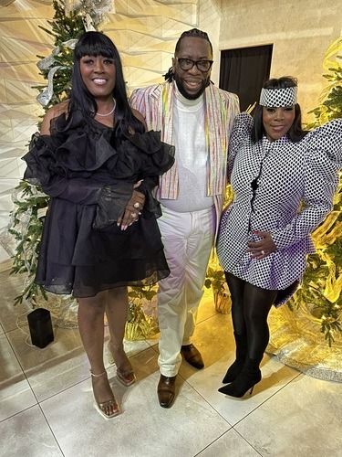 Three people posing indoors at a formal event, dressed in black, white, and gold outfits.