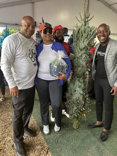 Four people posing indoors beside a decorated Christmas tree, one holding a gift basket.