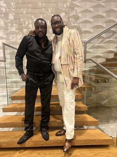 Two men posing on a staircase, one in black and one in a cream striped suit, smiling indoors