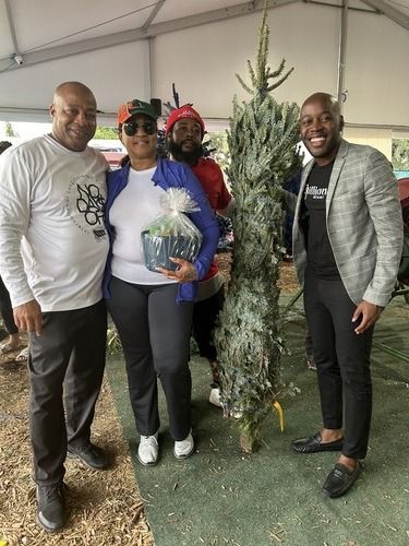 Four people stand together indoors, smiling and holding a wrapped gift beside a decorated Christmas tree.