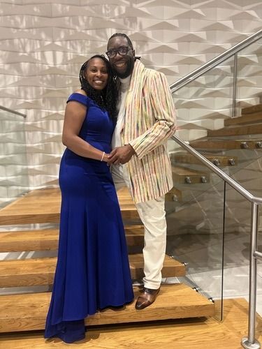 Two people smiling and hugging on a staircase, with a glass railing and textured wall behind them.