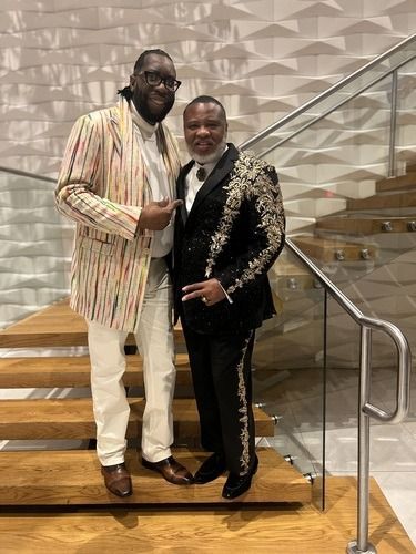 Two people posing on stairs, one in a striped jacket and one in a black sequined suit, smiling.