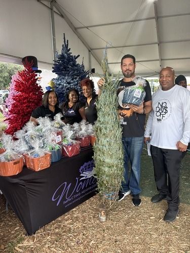 Group posing at a holiday market booth with decorated trees, gift baskets, and a “Merry Christmas” display.