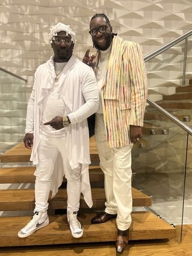 Two men posing on stairs, both in white and cream outfits, one holding a drink and smiling.