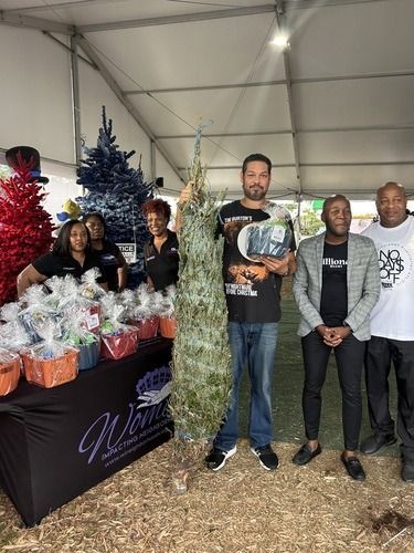People posing at a holiday market booth with Christmas trees and wrapped gift baskets under a tent