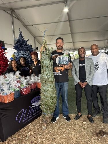 People standing at a holiday market booth with Christmas trees and gift baskets inside a tent