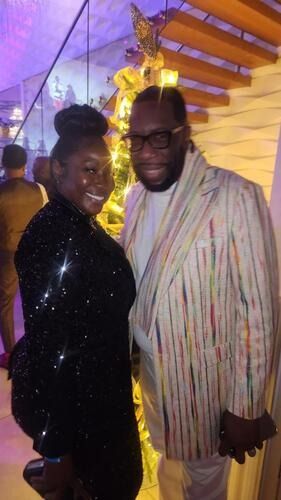 Two people smiling at an indoor event, one in a black sequined outfit and the other in a striped shirt.