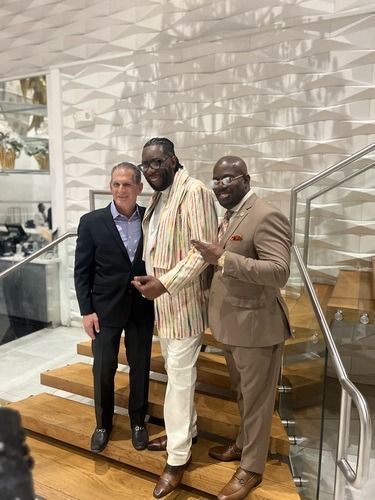Three men posing on indoor stairs, one in a black suit, one in a striped shirt, one in a tan suit.