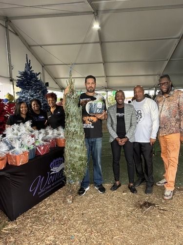 Five people pose beside a festive holiday display inside a tent, with decorated trees and wrapped gifts.