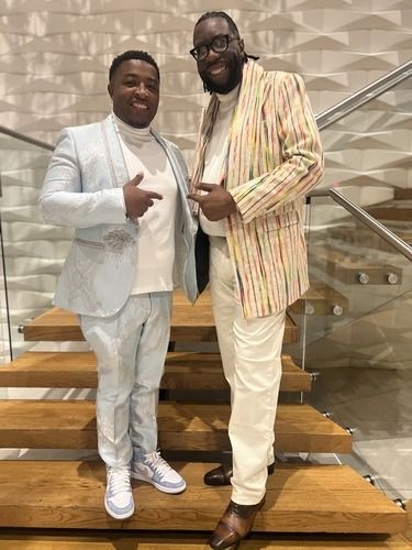 Two men posing on indoor stairs, wearing light-colored suits and smiling at the camera.