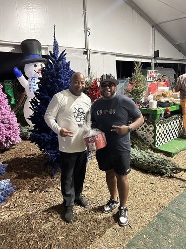 Two people posing at a holiday display with a snowman and decorated trees, holding a gift bag.