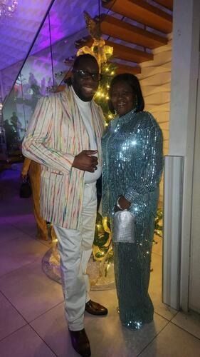 Two people posing indoors at a festive event, wearing striped and sparkling outfits under purple lighting.