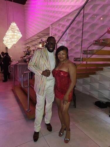 Two people posing at a pink-lit event, with stairs and a chandelier in the background.
