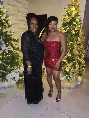 Two people posing in formal dresses beside lit holiday trees.