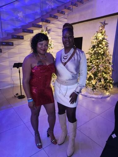 Two women posing at a holiday party beside a lit Christmas tree and staircase.