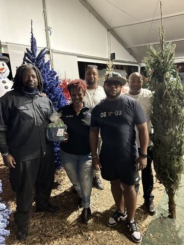 Four people standing outdoors at a holiday tree lot, posing beside decorated Christmas trees.