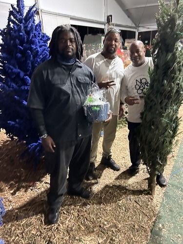 Three people standing outdoors by decorated trees, one holding a gift basket at night.
