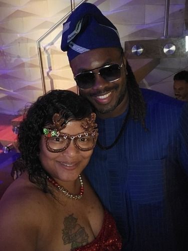 Two people smiling and posing together indoors, one in a blue outfit and headwrap, the other in a red dress and glasses.