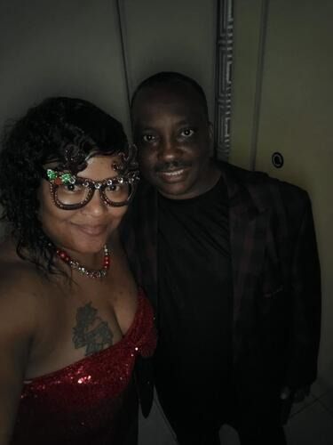 Two people smiling together indoors, one in a red sequined dress and glasses.
