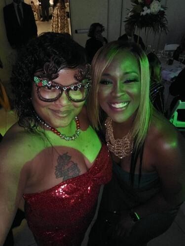 Two people smiling at a dimly lit event, one in a red sequined dress and one in a black dress.