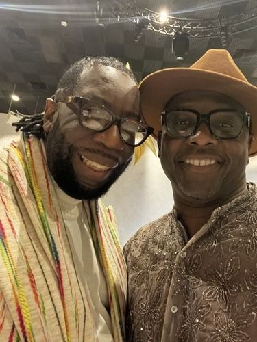 Two men smiling for a selfie indoors, one in glasses and a cream scarf, the other in a tan hat and patterned shirt.