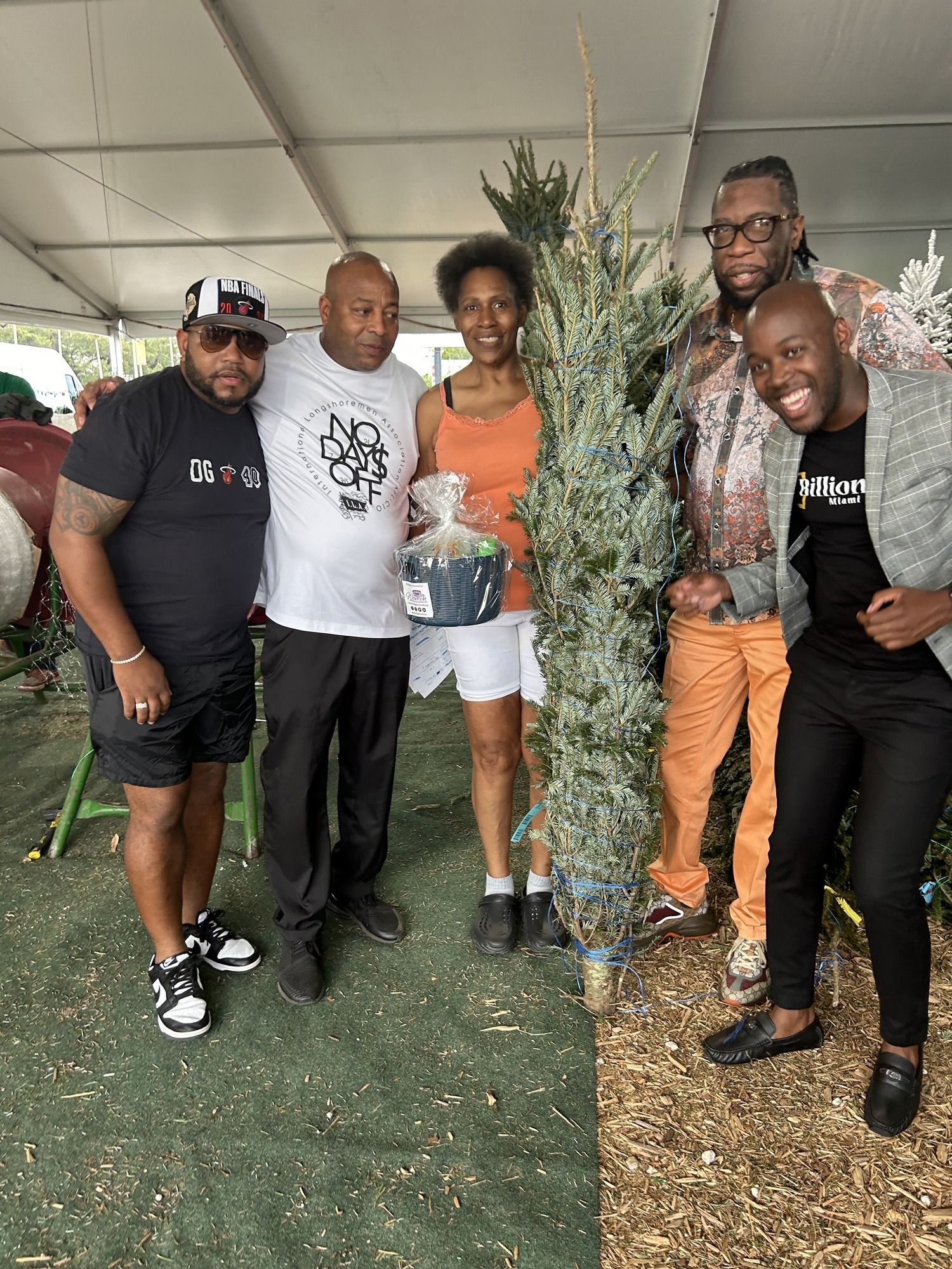 Five people posing together under a tent, with one person partly hidden behind a camouflage tree prop.