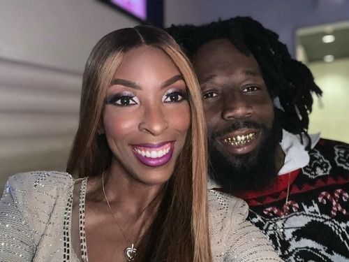 Two smiling people posing together indoors, one in a sparkly silver top and one in a patterned sweater.