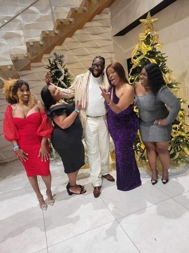 Five adults posing together at an indoor celebration, with decorated Christmas trees in the background.