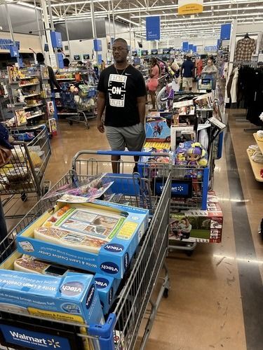 Man standing in a Walmart aisle behind a shopping cart filled with boxed items and snacks.