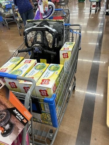 Shopping cart filled with boxed goods in a supermarket aisle, with a black bag on top.