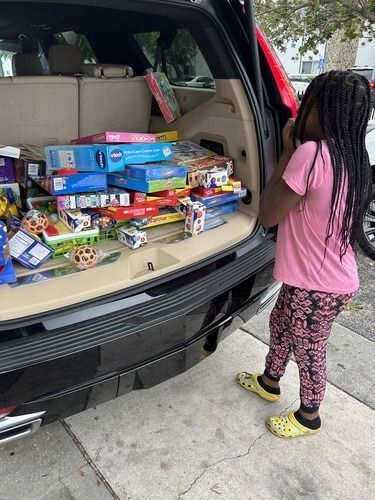 Open SUV trunk packed with toy and game donations; child stands beside it.