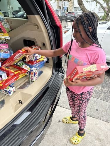Girl in pink shirt and patterned leggings reaching for snacks from a car trunk filled with groceries.