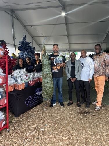 Group of people standing at a holiday market booth with gift baskets and a decorated tree under a white tent