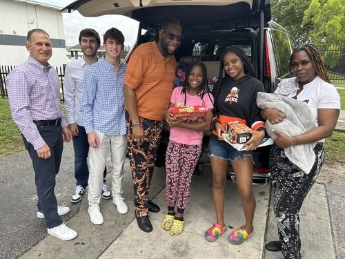 Six people stand together at an open van, holding bags and a blanket, smiling for a group photo.