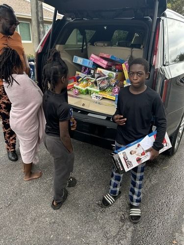 Three children load boxes into the open trunk of a red SUV on a street.
