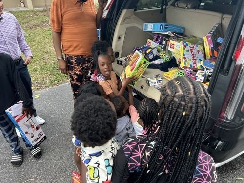 Children receiving toys from the back of a van on a street, with boxes stacked inside.